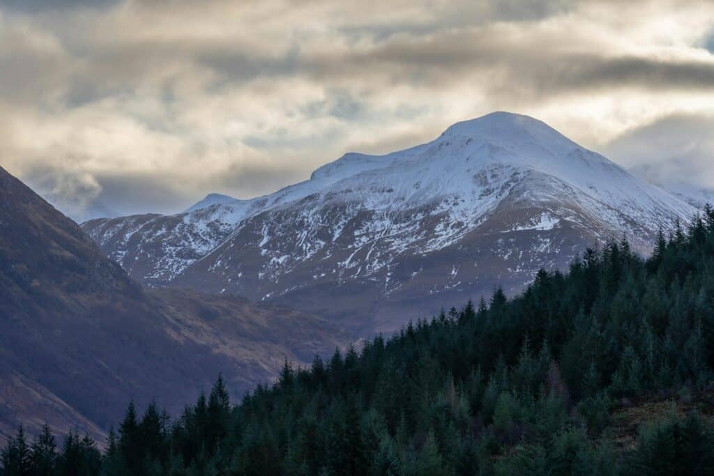 Breathtaking view of snow-capped Ben Nevis, Scotland, enveloped by clouds and forested hills.