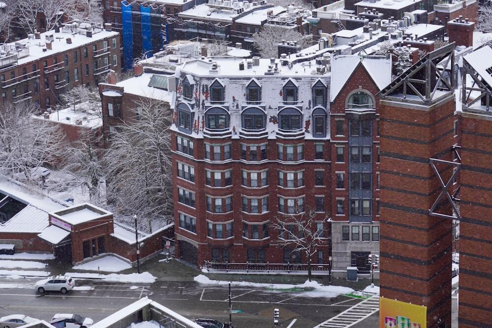 A historic Boston brownstone covered in fresh snow, capturing the charm of the city’s Beacon Hill and West End architecture. The intricate detailing of the dormer windows and red brick facade contrast beautifully against the winter backdrop, offering a glimpse into Boston’s rich architectural heritage. Snow-covered trees and rooftops add to the serene winter atmosphere of this classic New England setting.