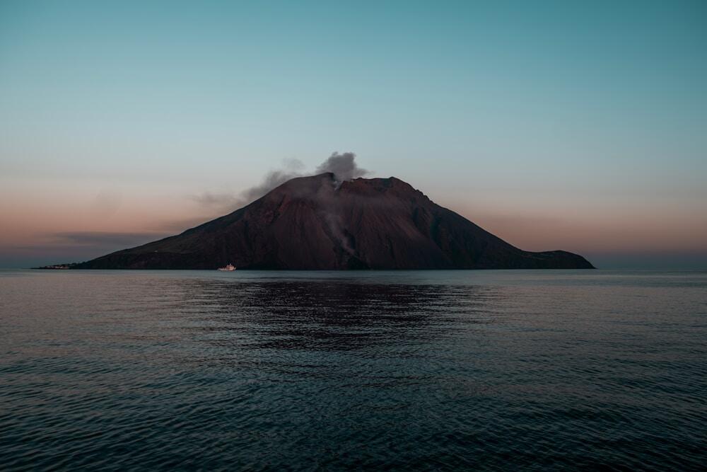 A breathtaking view of Stromboli, one of the most active volcanoes in the world, captured at dusk. The island stands majestically against the serene sea, with gentle plumes of smoke rising from its crater, showcasing the raw power of nature.